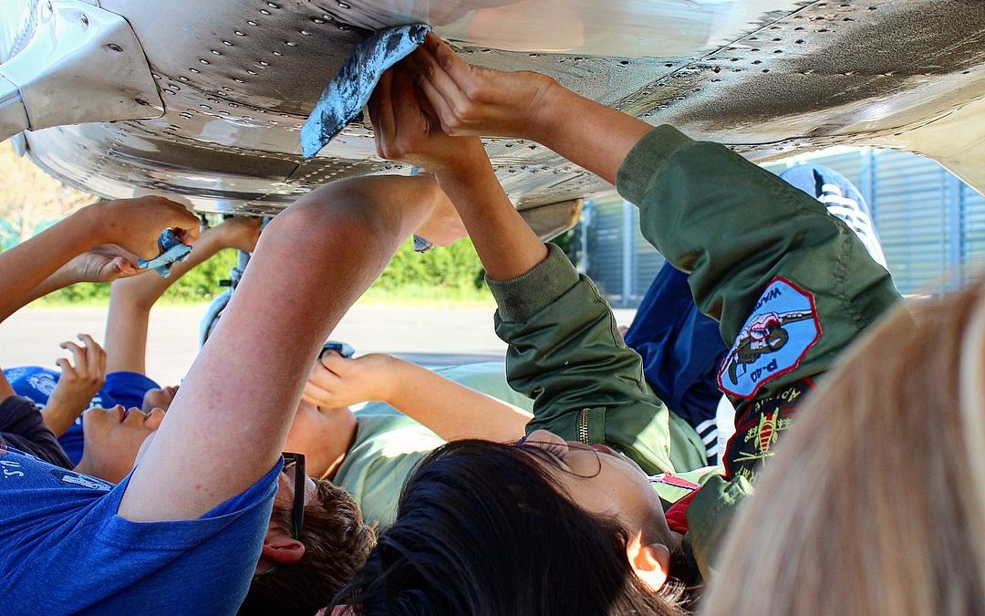 Aviation explorer members are lying on their back under and airplane as they gently remove oil and dirt during an airplane wash fundraiser.